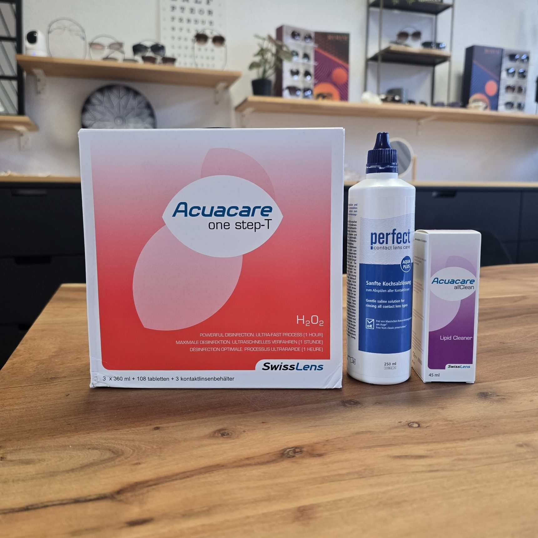 Pflegemittel für Sklerallinsen: Acuacare One Step-T Peroxid-System, Perfect sanfte Kochsalzlösung und Acuacare allClean Reiniger auf einem Holztisch in einem modernen Optikerladen.